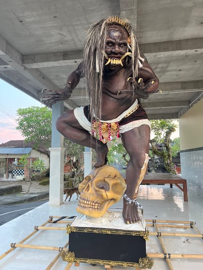 This image captures a large effigy, consistent with an Ogoh-Ogoh, situated in Celuk, Indonesia, beneath a concrete structural overhang. The figure has dark skin, long, stringy white and grey hair, a detailed face with prominent fangs, sharp teeth, and elongated yellow fingernails on its clawed hands. It is depicted in a crouching pose, with its right foot resting on a large, golden human skull. The left ankle features a black and white checkered fabric tie and a gold chain. The effigy wears a white and dark brown loincloth-style garment adorned with elaborate gold, red, and white decorative elements around the waist, and a feathered headpiece. It rests on an ornate black pedestal with gold accents, which is supported by a visible bamboo frame on a white tiled ground surface. In the background, a street is visible with residential structures featuring tiled roofs, a building with a black and white checkered facade, green trees, and utility lines against a sky showing pink and blue tones. A small brown dog walks near a white pillar in the mid-ground. To the right, a wooden bench and additional trees are present.