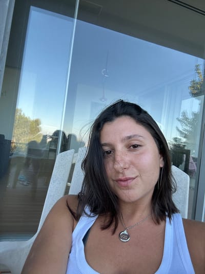 A female subject is depicted in a mid-shot, situated in the city of Merom HaGalil Regional Council, Israel. She has dark, shoulder-length hair and a complexion with visible freckles across her nose and cheeks. She is wearing a white ribbed tank top and a silver-toned necklace featuring a circular pendant with an indiscernible emblem. A single long, thin silver-toned dangle earring is visible on her left ear. She is seated in a light-colored, slatted outdoor chair, consistent with a patio or Adirondack style, and is looking directly towards the camera with a slight, neutral expression. Behind her, a large glass panel, likely part of a sliding door or window, reflects a clear blue sky, green foliage suggesting an outdoor landscape, and elements of an interior space including a ceiling and a light fixture. The scene is illuminated by natural daylight, indicating an outdoor patio or balcony setting.