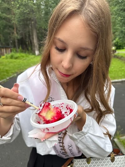 A young person with long brown hair is about to eat a candy apple. They are wearing a white blouse and a pink sash that reads "Kucera Bboy". The candy apple is red and white, with sprinkles, and is on a stick. The person is holding the candy apple and looking down at it. In the background, there is greenery, suggesting an outdoor setting. The lighting indicates it is daytime. There is a designer bag to the right of the frame.