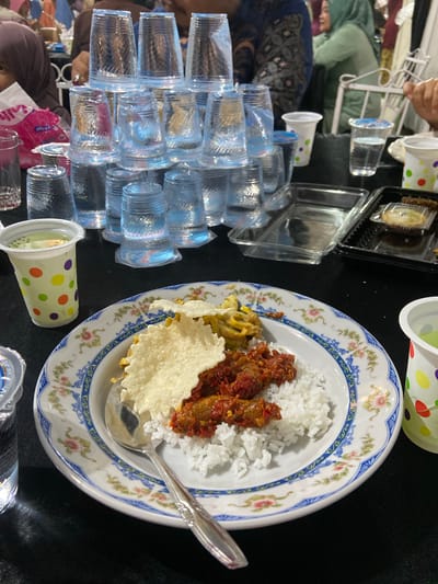 The image depicts a table laden with food and drinks, likely during a meal or gathering in Air Molek II, Indonesia. The scene includes stacks of clear plastic cups, some filled with water and others empty. One prominent object is a white plate with a decorative blue and floral rim, filled with white rice, a reddish-brown dish likely containing meat in sauce, and a piece of crispy cracker or wafer. A silver spoon rests on the plate. To the left, there are several clear plastic containers, one of which holds what appears to be a dessert or pastry. In the background, partially visible, are people. One person is wearing a light green headscarf and a dark top, and another is wearing a dark blue headscarf with a patterned fabric. The setting appears to be indoors, with a dark tablecloth covering the table. There are also some colorful paper cups with polka dot patterns. The lighting suggests it could be daytime or artificial lighting, but no specific time of day or weather can be determined. No discernible text is visible in the image, apart from what might be branding on a pink plastic bag in the lower left corner, which is too blurry to read. The overall impression is one of a communal meal, possibly a celebration or a regular gathering.