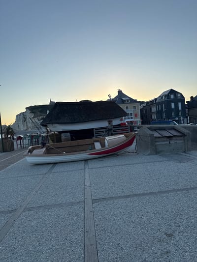 A small, white and red rowboat rests on a paved walkway near the coast in Étretat, France. The boat is angled slightly towards the viewer, with its bow facing left. Behind the boat, a building with a thatched roof sits on stilts, and several tall masts are visible. Further back, a line of buildings with dark roofs and light-colored facades stretches along the waterfront. The sky above is a pale blue, transitioning to a soft yellow at the horizon, indicating either sunrise or sunset. In the distance, dramatic white cliffs rise from the sea. The walkway is made of grey paving stones with wooden strips embedded in a geometric pattern. A concrete structure with a wooden lid is also present on the walkway to the right of the boat.