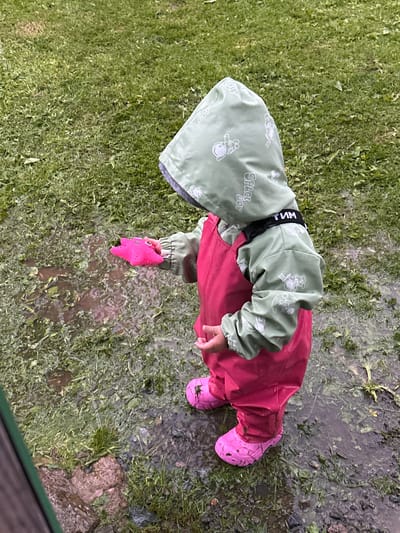 A young child, wearing a light green hooded rain jacket with a pattern of cartoon astronauts and the words "SPACE BOY," and pink overalls, stands in a puddle of water on a grassy area. The child is also wearing pink boots. In their right hand, the child holds a bright pink, star-shaped toy. The environment appears to be outdoors, with cut grass and puddles indicating recent rain. The scene is devoid of any urban elements typically associated with a city, suggesting it is likely a rural or park-like setting. The location specified as Азинский, Russia, cannot be confirmed or denied by visual cues in the image.
