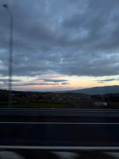 The image displays a landscape scene viewed through a window, captured during either sunrise or sunset. The perspective suggests the photograph was taken from inside a structure, possibly a building or a vehicle, looking outwards.
The upper portion of the image is dominated by a vast and dramatic sky, characterized by heavy, dark grey and blue-grey clouds. These clouds are thick and appear somewhat turbulent. Towards the middle and right side of the sky, the clouds thin or break, revealing a strip of softer light. This area exhibits a beautiful blend of pale yellow, soft orange, and subtle pink hues, indicative of the sun being low on the horizon, either rising or setting. Smaller, more defined cloud formations are also visible within this lighter band of sky.
Below the horizon, the landscape unfolds with gently rolling hills and mountains. The foreground and mid-ground hills are covered in lush green vegetation, possibly agricultural fields or pastures, and are dotted with clusters of small buildings or houses, suggesting a rural village or settlement. Further in the distance, mountain ranges appear darker and more subdued in color, partially obscured by the ambient light conditions and distance.
On the far right side of the image, a prominent dark, vertical band extends from top to bottom, which is clearly a window frame. This frame, along with a narrower vertical line to its left, creates a visual barrier, framing the landscape view. There are also hints of reflections or internal structures within the dark frame. A faint, translucent, elongated, light-colored rectangular shape, possibly a reflection or a part of the window's structure, extends diagonally from the lower left corner towards the center-left sky. Minor smudges or streaks are also discernible on the window's surface, particularly in the lower-left area.
The overall mood of the image is serene yet somewhat dramatic, conveyed by the contrasting light and dark elements of the sky and landscape. No text is visible in the image.