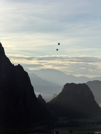 This is a vertical, outdoor photograph captured from a high vantage point overlooking a valley in Naka, Laos. The foreground is dominated by the dark, silhouetted mass of a steep, rocky mountain on the left side of the frame. To the right, a similarly shaped, but smaller, dark mountain rises, with a more rounded, forested peak.

Beyond these foreground mountains, a series of progressively lighter, mist-shrouded mountain ranges recede into the distance, creating a sense of depth. These ranges appear layered, with a hazy, atmospheric perspective softening their details.

In the sky, high above the mountains, three hot air balloons are visible. They are dark, teardrop shapes against the pale, cloudy sky. The balloons are positioned in a diagonal line, with the uppermost balloon to the right of the other two.

The sky itself is a pale blue, with horizontal bands of white and light gray clouds, some of which are illuminated by the sun from behind, creating a subtle glow.

At the base of the mountains, in the valley, hints of human habitation can be discerned, including what appear to be agricultural fields, possibly rice paddies, and small structures. The overall atmosphere of the image is serene and expansive, suggesting an early morning or late afternoon light.