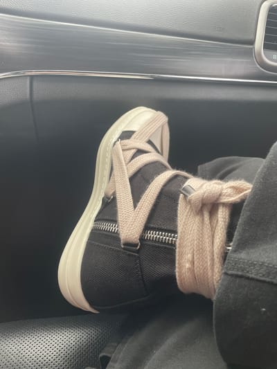 A close-up, eye-level shot shows a person's foot wearing a black high-top sneaker with a thick, light-tan lace that has been tied around the ankle. The sneaker features a white sole, a zipper detail on the side, and metal eyelets. The person is wearing black pants. The background consists of the interior of a vehicle, with a black dashboard and a metallic trim piece visible. The image appears to have been taken while the person is a passenger in a car, possibly in Cuautitlán Izcalli, Mexico, given the context provided.