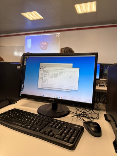 This image depicts a computer laboratory or classroom located in Gandra, Portugal, likely within an educational institution specializing in health or dental sciences.
In the immediate foreground, a workstation is visible with a black Philips monitor displaying a blue desktop background. Two overlapping file explorer windows are open on the monitor; the front-most window is titled "Scan File Import" and shows a directory path containing "Gandra," indicating the local file system. A black Logitech keyboard is positioned directly in front of the monitor, with a black Logitech mouse to its right. Black cables are clustered between the monitor and mouse. Dark-colored computer towers are partially visible on either side of the monitor.
The environment features light-colored walls, with a red pipe running horizontally across the upper portion of the back wall. Two rectangular fluorescent light fixtures are mounted on the white ceiling.
In the midground, the backs of several individuals' heads are discernible, indicating other students or participants seated at adjacent workstations.
The background reveals a large white projection screen on the far wall. A projector displays a detailed 3D model of a human molar tooth onto this screen, suggesting an instruction or demonstration session related to dental design or computer-aided design/manufacturing (CAD/CAM) in a dentistry or dental technology program.