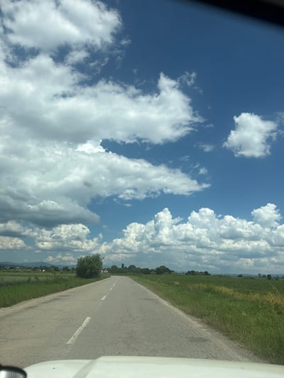 The image captures a paved road in Musachevo, Bulgaria, leading through a rural landscape under a dynamic sky filled with cumulus clouds. The road, marked with a white dashed line, is bordered by lush green fields on both sides. In the distance, trees and a few houses are visible, with rolling hills forming the horizon. The overall scene depicts a serene, daytime view of the Bulgarian countryside.