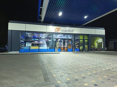 The image shows a nighttime exterior view of a "Smarket" convenience store located in the city of Guliston, Tajikistan. The store is part of a larger structure with a blue ribbed ceiling and white trim, illuminated by overhead lights. The storefront features large glass windows displaying various products and shelves. Prominently displayed on the building's facade is the "Smarket" logo. To the right of the main entrance, there is a large advertisement featuring a cup of coffee with the text "S coffee." A red and black bicycle is parked in front of this advertisement. The foreground consists of a paved area with patterned stone tiles and asphalt. The scene is deserted.