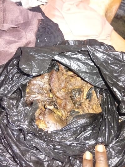 A black plastic bag containing cooked meat and bones sits on a surface. Two fingers are visible in the bottom right of the frame. The background includes fabric in various colors. The location is Aviation Quarters, Nigeria.