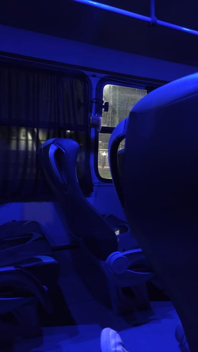 The image displays the interior of a passenger bus under predominant blue artificial lighting, situated in La Tijera, Mexico. The scene is observed from a seated position looking forward. The floor in the lower frame is dark blue. A pair of feet wearing white athletic shoes is present in the lower right, indicating a passenger's position. The foreground and midground feature the backs of two modern, dark blue-grey passenger seats: one on the left and a larger, closer one on the right. A window on the left side is obscured by dark curtains. A rectangular window on the right reveals a dimly lit external environment, consistent with a nighttime setting, showing indistinct structures and possibly part of a fence. Through this window, the lower torso and legs of an individual wearing dark trousers and light-colored shoes are partially visible outside the vehicle. A small, light-colored device is mounted on the interior wall beneath the right window. An overhead handrail is visible in the upper portion of the frame.