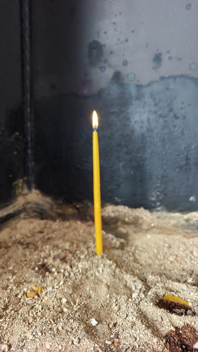 A single yellow candle burns brightly in a sand-filled holder. The candle is positioned vertically in the center of the frame, its flame a vibrant point of light. The sand appears to be composed of fine grains mixed with small pebbles. In the foreground, scattered remnants of what appear to be burnt offerings or debris are visible. The background consists of a dark, rough-textured surface, possibly a wall or the interior of a structure. The overall impression is one of a solemn or religious setting, potentially within the city of Selçuk, Turkey, known for its historical and religious sites. There are no people or animals visible in the image.