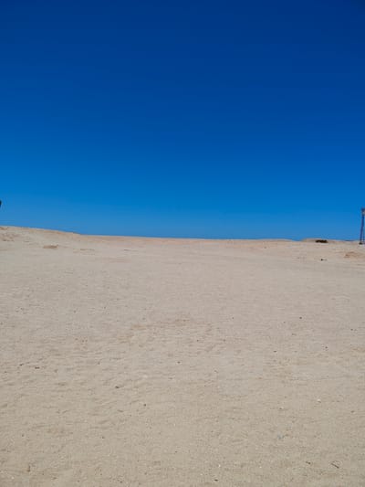 The image depicts a vast, sandy expanse under a clear, brilliant blue sky. The ground is a light beige, textured with fine grains of sand and occasional small pebbles or debris. The terrain appears relatively flat, with a gentle rise towards the horizon. In the distance, the horizon line is clearly defined, separating the sandy ground from the unbroken blue of the sky.

On the far right of the image, a tall, dark, lattice-like structure resembling a telecommunications tower or pylon stands against the sky. On the far left, a very small, dark, indistinct shape can be seen, possibly a person or an object, but it is too far away and out of focus to be identified clearly. There are no other discernible subjects or significant objects in the scene.

The scene suggests a remote, arid environment, likely a desert or a desolate coastal area. The bright blue sky and the absence of clouds indicate fair weather. The strong, even lighting across the sand suggests it is midday or early afternoon, with the sun high in the sky. The overall impression is one of openness, stillness, and solitude. There is no visible text in the image.