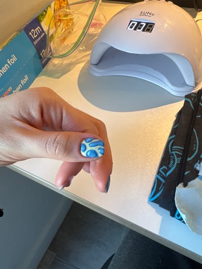 Here's a detailed description of the image:

A person's hand, specifically the thumb, is prominently displayed with a nail painted in a blue and white amoeba-like pattern.  The other fingers are visible, with what appears to be dark colored or clear nail polish.

Surrounding the hand, various objects are situated on what looks like a light-colored surface. To the left, there's a white UV nail lamp with a digital display showing "888" and the brand "SUN".  Beneath the lamp is a blue box labeled "SPAR" and "chen Foil" with the text "12m 300mm wide". A transparent plastic bag or wrap is also visible.

In the background, there is a black and blue printed fabric or object. The surface upon which the objects are placed seems to be a light-colored table or countertop. The right-hand side has a view of a gray surface, possibly part of the room's floor or another table.

The location within the city of Hull, United Kingdom, cannot be determined from the image.