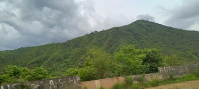 The image depicts a lush green mountainous landscape under a cloudy sky. The slopes are covered with dense vegetation, with trees scattered across them. The foreground features a concrete barrier with graffiti on it. The scene appears to be a natural environment, possibly a rural or park area within the city of Gu, Nigeria. The weather seems overcast, contributing to a somewhat muted color palette. No people or specific actions are discernible in the image.