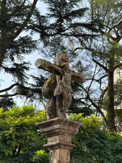 A stone sculpture depicting Jesus Christ on the cross stands on a pedestal in Santiago de Compostela, Spain. The sculpture is weathered and covered in moss. Behind the sculpture, lush green foliage and tall trees with dark branches and needles create a natural backdrop against a pale sky. A portion of a building with a gray, sloped roof is visible in the upper right corner of the image.