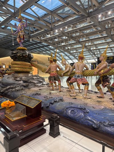 This is a vertical, full-shot photograph of a large, elaborate sculpture depicting figures from Thai mythology, likely located in a public indoor space, such as an airport terminal in Nong Prue Subdistrict, Thailand. The sculpture is vibrantly colored and intricately detailed, featuring several human-like figures wearing traditional Thai headdresses and ornate clothing. These figures appear to be engaged in some form of struggle or ceremonial act, as they are posed dynamically and connected by a large, serpentine creature.
The sculpture is set against a backdrop of what appears to be a stylized representation of water and land, with various textures and shades of blue, brown, and earth tones. The figures themselves are sculpted with realistic human anatomy and adorned with gold jewelry, adding to the richness of the artwork.
Above and behind the sculpture, the architecture of the interior space is visible, characterized by a large, modern glass and steel ceiling structure, indicative of a contemporary building. The lighting suggests it is daytime, with natural light filtering through the expansive windows, supplemented by interior lighting.
Visible text in the image includes what appears to be airport gate information ("D1-8", "- D8"), airline logos ("QATAR"), and possibly signage in Thai and Chinese, reinforcing the context of an international transportation hub. The overall impression is one of cultural artistry and grandeur, presented in a busy, well-lit public environment. The presence of an offering of orange marigold flowers in the foreground, placed near a display case, suggests a cultural or religious significance to the sculpture.