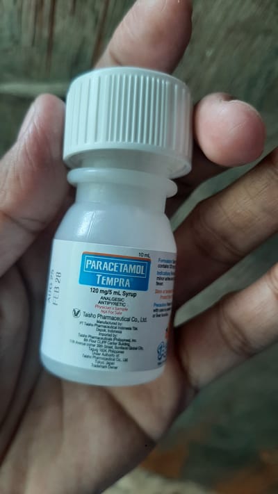 A hand holds a small, white plastic bottle with a blue and white label. The bottle contains liquid and has a ribbed white cap. The label is partially visible, showing the brand name "PARACETAMOL TEMPRÅ" and "120 mg/5 mL Syrup". Text on the label indicates it is an analgesic and antipyretic, a physician's sample, and "Not For Sale". Manufacturing and importing information is also present, including "Taisho Pharmaceutical Co., Ltd." and locations in Indonesia and the Philippines. The expiration date "AUG 25 FEB 28" is visible on the side of the bottle. The background is blurred and appears to be a wooden surface. The scene is indoors, with the bottle being the primary subject. There is no discernible context to indicate the location within the city of Kawit, Philippines.