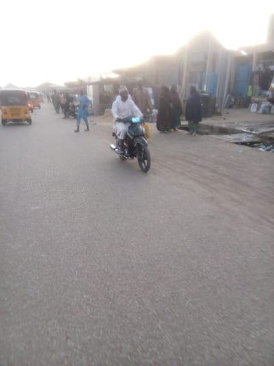 The image captures an outdoor street scene in Tudun Murtala, Nigeria, likely during early morning or late afternoon due to the soft, bright, and slightly hazy light. The central focus is a man in a white traditional robe and cap riding a black motorcycle on an asphalt road, heading towards the left side of the frame. The motorcycle's blue headlight is illuminated.
On the left side of the road, a yellow Keke Napep (auto-rickshaw) is visible, with another similar vehicle further down the road, indicating traffic or public transport activity. Several people are seen walking or standing along the roadside, including a man in a light blue garment on the left and women in dark traditional attire on the right.
The right side of the road features a row of simple structures, possibly market stalls or small shops, constructed from corrugated metal and other basic materials. The ground immediately adjacent to these structures appears unpaved and somewhat uneven, with some debris visible. The overall setting depicts a busy local thoroughfare or market area. No visible text is discernible in the image.
