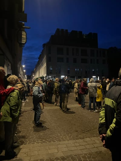 The image depicts a nighttime street scene in Rouen, France, specifically on Rue du Gros Horloge, identified by the prominent "GRANDE PHARMACIE DU CENTRE" sign on the left. Approximately 50-70 individuals, primarily adults, are gathered on a cobblestone street, facing various directions. The sky is dark blue, indicative of twilight or early night, with artificial streetlights illuminating the scene and casting shadows.

On the left, a building displays an illuminated, ornamental sign for "GRANDE PHARMACIE DU CENTRE" and a dark green pharmacy cross symbol above it. Further down the street on the left, additional streetlights are visible. On the right side of the street, multiple buildings with illuminated windows line the path, including a modern, multi-story structure with a flat roofline in the mid-distance.

The people are dressed in casual, cool-weather attire, including various jackets and coats; one individual near the right-center wears a bright yellow raincoat. Several individuals are holding mobile phones, some pointed upwards or towards the street. Their collective stance suggests they are observing an event or phenomenon not visible within the frame. The cobblestone pavement is visible across the entire foreground and midground.