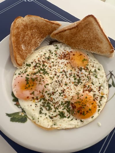A plate of fried eggs and toast sits on a blue and white striped placemat. The fried eggs are seasoned with parsley and red pepper flakes. The toast is golden brown and cut into triangles. The plate has a floral pattern. The scene is indoors, likely a home kitchen or dining room, in Sant Feliu de Guíxols, Spain.