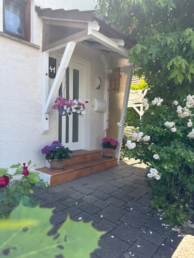 The image depicts the front entrance of a house with a white door and a small covered porch. The porch is supported by white wooden beams and has a white railing. A house number "41" is visible on the wall to the left of the door. There are several potted plants in front of the door, including a hanging basket with pink and white flowers and two potted hydrangeas. To the right of the entrance, there is a large bush with white roses. The ground in front of the entrance is paved with grey interlocking stones. The scene is set in a residential area, likely Bietigheim-Bissingen, Germany, given the architectural style and vegetation. The overall impression is one of a well-maintained and welcoming home exterior on a sunny day.