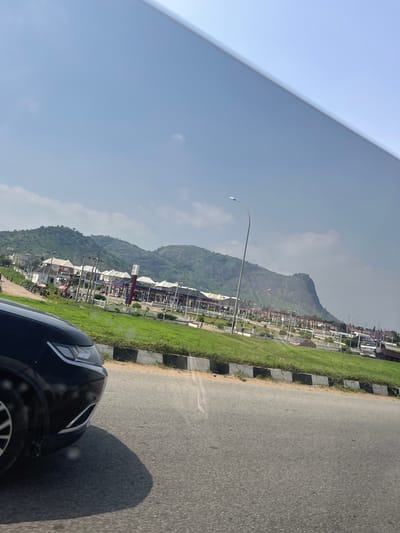 The image depicts a roadside scene in Sabojibi, Nigeria. The perspective is from within a vehicle, with the front of a dark-colored car visible in the foreground on the right. The road itself is asphalt, with a grassy median to the left, bordered by concrete blocks. Beyond the median, a line of buildings, likely shops or businesses, is visible. These structures have white roofs and some display red and blue signage. In the background, a large, green, mountainous landscape dominates the scene under a bright, clear sky. A tall lamppost is present on the grassy median. The overall impression is of a sunny day in a developed, possibly suburban or commercial area situated near a natural landscape. No people are clearly discernible.