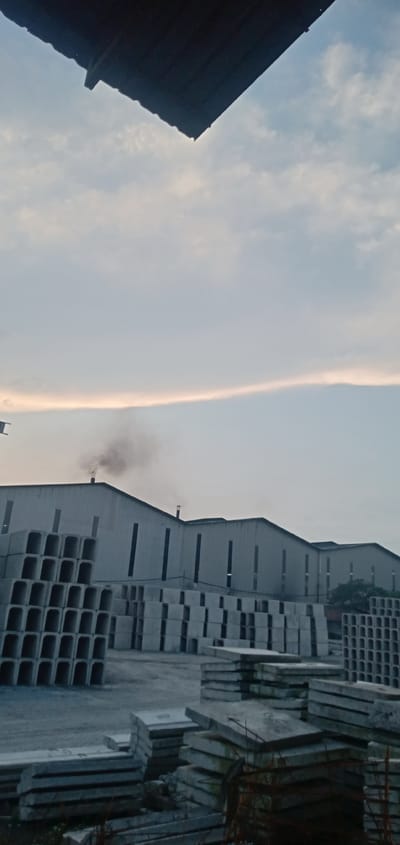 The image depicts an outdoor industrial area in Sumput, Indonesia. In the foreground are stacks of concrete slabs and rectangular concrete frames, likely precast construction elements. Behind these are rows of large, gray, warehouse-like buildings with vertical windows and pointed roofs. Smoke is visible emanating from one of the chimneys on the buildings. The sky is overcast with a light-colored horizon. A dark-colored roof edge is visible in the upper part of the image. The ground is mostly gray concrete or gravel. No people are visible.