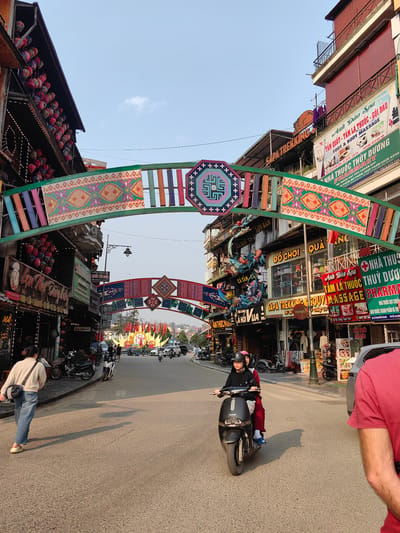 This is a daytime street scene in Sa Pa, Vietnam, characterized by a paved road flanked by multi-story commercial buildings. Two decorative arches, adorned with colorful fabric panels displaying traditional Hmong geometric patterns, span the street; the closer arch is green, while a second, similar arch is further down. The building on the left features multiple rows of red and pink lanterns hanging from its balconies.
In the mid-ground, a black JAKAR electric scooter, operated by a person in a black jacket and pink helmet, carries a passenger in red pants and a pink helmet; both are traveling towards the camera. On the left sidewalk, a person in a beige top and blue jeans walks away from the camera, passing several parked motorbikes. The immediate foreground on the right shows the partial back view of a person wearing a red t-shirt.
Numerous motorbikes are parked along both sidewalks, and other vehicles, including cars and additional motorbikes, are visible in the distance. Businesses lining the street include "SAPA TREKKING," "ANH ĐÀO SPA TẮM LÁ THUỐC - GỘI ĐẦU FOOT & BODY MASSAGE," "NHÀ THUỐC THÙY DƯƠNG PHARMACY," and "ĐỒ CHƠI - QUÀ" (Toys - Gifts). A large, festive-looking display with flags is visible further down the street. The sky is clear, and shadows are cast across the pavement by the sunlight.