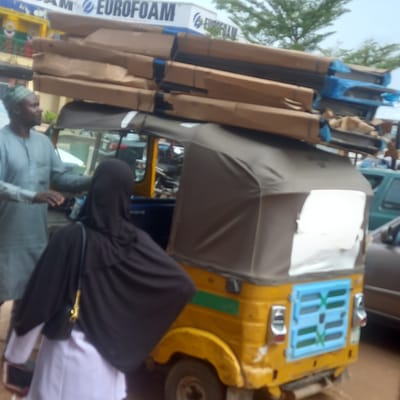 A yellow motorized tricycle (auto-rickshaw) is heavily loaded with stacked rectangular packages marked "EUROFOAM." The vehicle is parked on a city street in Sardauna, Nigeria. An adult male, wearing a white shirt and cap, stands beside the auto-rickshaw. Another individual, draped in a black hijab, is positioned next to the auto-rickshaw. The scene suggests a commercial transaction involving the transportation of goods.