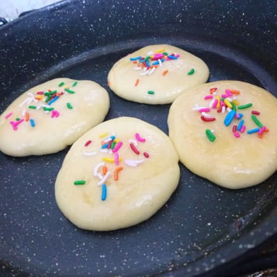 Four light-colored, circular pastries topped with colorful sprinkles are arranged in a non-stick frying pan. The pastries appear to be raw dough or partially cooked. The frying pan has a dark, speckled surface. The background is out of focus but shows a white countertop. There are no people visible in the image. The scene is a close-up shot, focusing on the pastries and the pan. There is no indication of the location being El Limón, Venezuela.