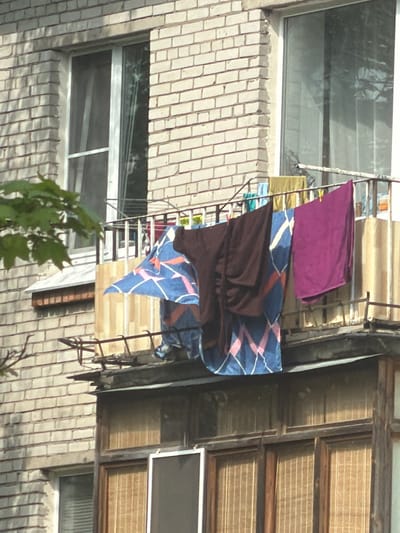 The image depicts an exterior view of a multi-story brick building, possibly an apartment complex, in Tokkari-Lend, Russia. Clothes are drying on a metal rack extending from a window. The laundry includes a purple towel, a patterned blue and brown blanket, and other unidentified items. Windows are visible on the brick facade. The building features both old wooden-framed windows with blinds and more modern windows with metal frames. Green foliage is visible in the foreground.