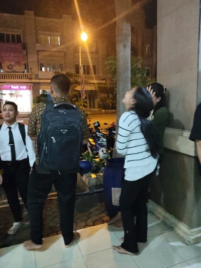The image shows three people standing outside at night in Telukjambe, Indonesia, located at the coordinates -6.308 latitude and 107.282 longitude.  Two men and a woman are standing near a building with a large blue trash can nearby. One man is wearing a white shirt, black pants, and a black tie. He is standing on the left side of the image with his hands at his sides. The second man, wearing a black backpack, black pants, and a floral shirt, is standing in the middle of the image. The woman is wearing a white and black striped long-sleeved shirt and black pants. She is standing on the right side of the image, looking up and smiling. She has long black hair. The background of the image is blurry, but it shows a building with a light on top of it. The scene is well-lit, though the source of light is unclear.  The image shows a casual nighttime interaction between these three individuals.