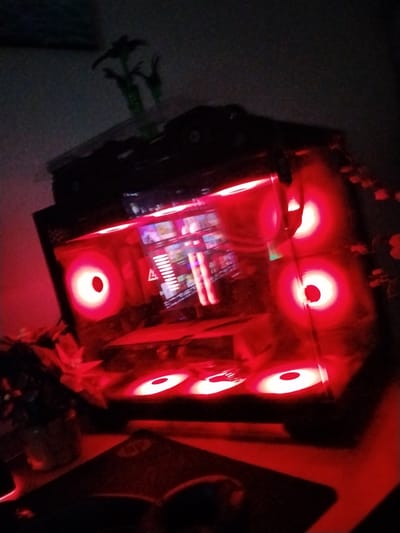 The image shows a gaming computer case with red LED lighting. The computer case is placed on a desk in a dimly lit room. There are also some plants and a mousepad visible on the desk. The image appears to be taken at night. The location is specified as the city of Holon, Israel. There are no people or actions depicted in the image. The scene is static and focuses on the computer setup.