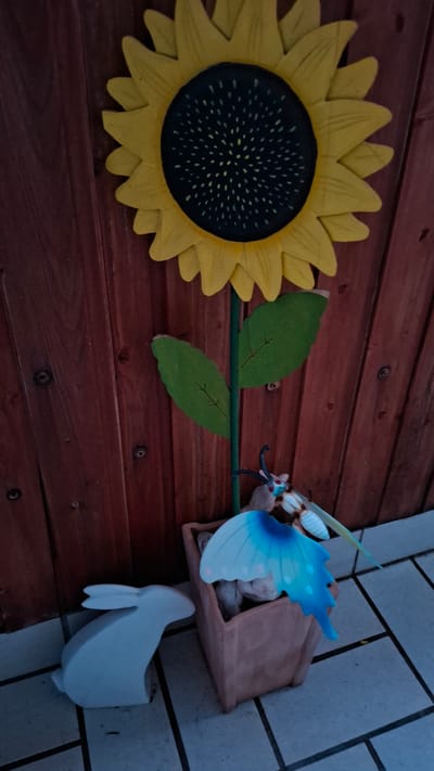 A decorative metal sunflower is attached to a wooden fence. A butterfly and a white rabbit figurine are placed in front of the sunflower. The scene appears to be outdoors on a tiled surface, possibly a patio or walkway. The lighting suggests it is evening or a shaded area. There are no people visible in the image. The overall setting is a garden or outdoor decorative display. The location is not identifiable as Cologne, Germany.