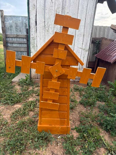 The image shows a wooden structure, possibly a birdhouse or a decorative garden piece, made of light-brown stained wood. It is placed outdoors on the ground, with dry soil and green weeds around it. In the background, there is a wooden fence or wall made of weathered, light-colored planks, and a glimpse of a metal roof. The structure itself is complex, with multiple wooden slats and panels arranged in a design that resembles a building with a peaked roof and several smaller, protruding elements. Some of these elements appear to be small signs or decorative pieces. The lighting suggests it is daytime, likely overcast or late afternoon due to the diffused light and shadows. The scene appears to be in a rural or semi-rural setting, possibly a backyard or a garden area, consistent with the location context of Ильчимбетово, Russia. There is no visible text in the image.