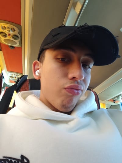 A young man wears a black, distressed baseball cap and a white hoodie. He has an earbud in his left ear. His expression is ambiguous, with slightly pursed lips. The background suggests he is on a bus, with overhead compartments visible and orange-colored paneling. A seatbelt is visible across his chest.