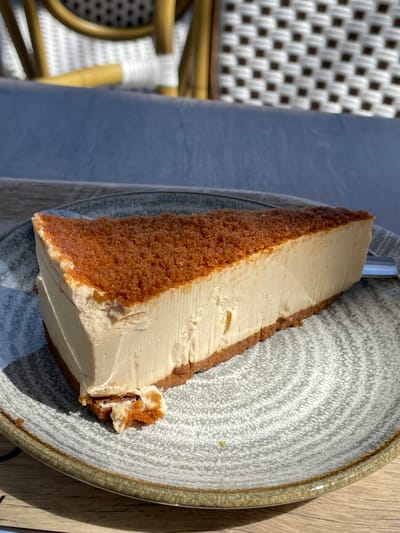A triangular slice of cheesecake with a crumbly brown topping and a graham cracker crust sits on a grey, patterned ceramic plate. The cheesecake is a light beige color, with some visible texture from the filling. A small piece has broken off the front edge of the crust. The plate rests on a wooden table, and in the background, a woven chair with a white seat and yellow frame is visible. The scene is lit by sunlight, casting shadows. This image was likely taken at an outdoor cafe in Algeciras, Spain, given the characteristic Mediterranean sunlight and the outdoor seating often found in such establishments.