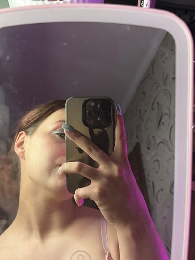 An unidentifiable person is taking a selfie in a mirror. The person's face is partially obscured by a smartphone held in their hand. The background appears to be a wall with textured wallpaper. The lighting is dim and a purplish hue is present, suggesting artificial lighting. The image offers no information about the location within Bishkek, Kyrgyzstan.