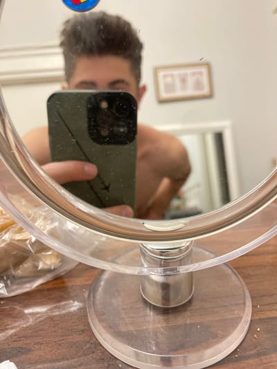 This is a close-up, waist-up mirror reflection of a young male with dark, styled hair. He is holding a dark green smartphone to take the photo. The background is blurred, showing a wall with what appears to be a framed picture with three rectangular images. The mirror is round and has a clear plastic stand. To the left of the mirror, a white plastic bag containing food items is visible on a wooden surface. The surface is cluttered with white powder. The setting is indoors, and there is no specific indication of the city of Bydgoszcz, Poland, within the image itself.