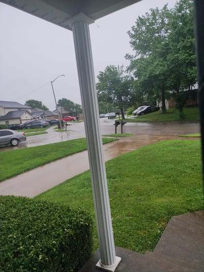 This is a medium-shot, vertical-orientation photograph taken from a porch, looking out at a street scene. The foreground features a white decorative column and the edge of a porch roof, obscuring the left side of the frame. The scene is dominated by a street lined with houses and parked cars. A paved sidewalk runs along the right side of the street, bordered by a lush green lawn. The weather appears to be overcast and rainy, as evidenced by the wet pavement and the dull sky. The foliage on the trees is dense and green, suggesting a summer or late spring setting.
Several cars are parked along the street, including a red pickup truck, a dark sedan, and a silver hatchback. In the middle distance, a black car is parked across the street from a house with a brick facade. The houses appear to be suburban residences. The overall mood of the image is somewhat somber due to the overcast weather. No people are visible in the frame. The time of day is indeterminate due to the lack of direct sunlight and shadows. The location is identified as Nicholasville, United States of America. No visible text is present in the image.