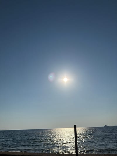The media file depicts a scenic view of the Adriatic Sea at Buljarica, Montenegro. The scene is dominated by a vast, clear blue sky that transitions to a lighter hue near the horizon. The sun is prominently positioned in the upper portion of the sky, emitting a bright glare with a subtle green lens flare visible below it.

To the right, the deep blue sea stretches out, its surface shimmering with countless reflections of sunlight, creating a dazzling, sparkling effect. Small waves are visible, indicating a gentle movement of the water. The shoreline is composed of pebbles, and a dark wooden post stands vertically in the water, partially obscured by the reflections. The overall impression is one of a serene and bright daytime at a coastal location. The image appears to be taken during daylight hours, likely late morning or early afternoon, given the intensity of the sun. The weather appears clear and sunny.
