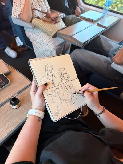 The image depicts a person sketching on a train. The individual, with dark nail polish and a white smartwatch, holds an open sketchbook with an ink pen. The sketchbook contains two line drawings of men's faces. To the left of the sketchbook, a small glass bottle of black ink sits on a wooden tray table. The train interior features a window with greenery visible outside, a folded tray table with a laptop and a water bottle, and seats occupied by other passengers. The scene is set in Rosny-sur-Seine, France, specifically on a train.