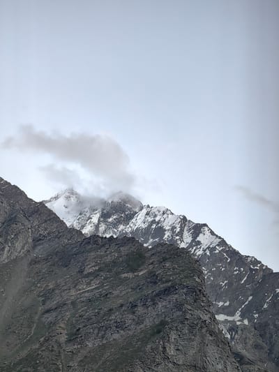 This image captures a majestic mountain landscape under a cloudy, overcast sky. The scene is dominated by rugged, rocky mountain slopes in the foreground, transitioning to snow-capped peaks in the background. The foreground mountains are dark and textured, showing layers of rock and sparse patches of greenery, suggesting a high-altitude, arid environment.
Further back, the mountains rise sharply, their upper reaches blanketed in white snow and ice, with dark rock faces peeking through. Wisps of clouds partially obscure the summits, adding a sense of mystery and depth to the scene. The sky is a uniform light gray, indicating a cloudy day, possibly with some atmospheric haze.
There are no people or objects visible, emphasizing the raw, natural beauty of the location. The overall impression is one of grandeur, solitude, and the imposing power of nature. The time of day is difficult to ascertain precisely due to the overcast conditions, but the muted light suggests it could be morning or late afternoon. The location is identified as Kead, India, a region known for its Himalayan topography, which aligns with the visual elements presented in the photograph. The absence of text further focuses attention on the natural landscape itself.
