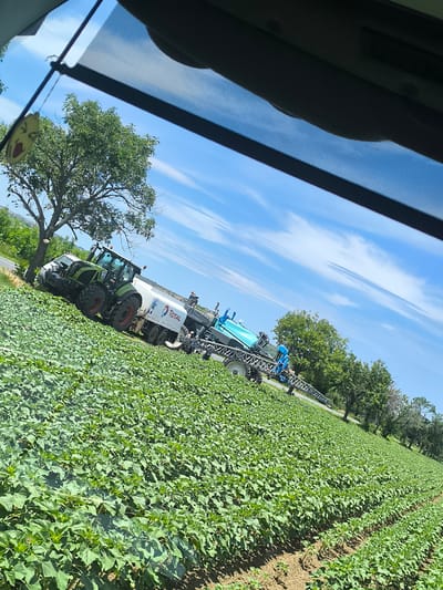 In a rural field in Inovo, Bulgaria, a green tractor pulls a white trailer with "TOTAL" branding, attached to a large blue agricultural sprayer. The tractor is positioned on the edge of a field filled with rows of young green plants, likely sunflowers given the location. Behind the tractor and sprayer, a line of trees borders the field. The sky is bright blue with wispy white clouds, indicating clear weather. The sun appears to be high in the sky, suggesting midday. The scene is captured from inside a vehicle, looking out through the windshield, with a portion of the vehicle's interior visible in the foreground. A yellow air freshener shaped like a smiley face hangs from the rearview mirror. The overall impression is of agricultural work in progress on a sunny day.