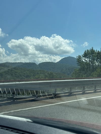 The image depicts a roadside view from a vehicle, likely on a highway, overlooking a mountainous landscape under a clear blue sky with scattered white clouds. In the foreground, a metallic guardrail runs horizontally across the frame, separating the road from the embankment. The road surface is asphalt with a white lane marker. The background features rolling hills covered in dense green foliage, characteristic of a forested mountainous region. Two tall, lattice-like towers, possibly power line pylons, are visible in the distance between the hills. A prominent, rounded mountain peak stands out further in the background, topped with a small structure. A large tree with green leaves is situated on the right side of the image. The overall scene conveys a sense of travel and natural scenery. The location is identified as the city of Razdolnoye, Russia.