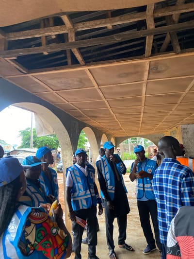 In Riyom, Nigeria, a group of individuals, many wearing matching blue caps and vests with reflective stripes, are gathered under a covered walkway. The ceiling of the walkway appears to be constructed of wooden beams and square panels, with some sections open to reveal the exposed roof structure. The individuals are of various ages and ethnicities, and some are engaged in conversation. One person is holding a piece of corn. The environment suggests an outdoor or semi-outdoor setting, possibly a market or a public gathering space, with glimpses of trees and buildings in the background. The overall atmosphere seems to be one of community engagement or a planned event.