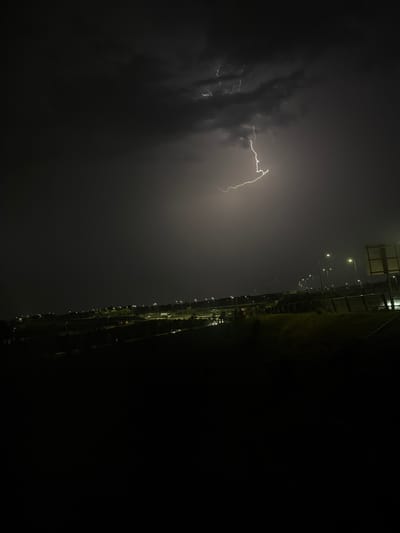 A nighttime scene captures a dramatic lightning strike illuminating dark, ominous storm clouds above a sprawling suburban landscape. The lightning forks across the sky, casting a bright, sharp light on the clouds and a portion of the horizon. Below, a dimly lit cityscape stretches out, characterized by rows of streetlights casting a warm glow on the empty roads and buildings. The foreground is dominated by dark, silhouetted hills and vegetation, suggesting a slightly elevated vantage point overlooking the illuminated area. The overall impression is one of a powerful natural event occurring over a quiet, unsuspecting urban environment. This appears to be a view of Badhoevedorp, Netherlands, given its characteristic flat terrain and the presence of numerous streetlights along what could be major roadways or developed areas. There are no people or discernible human activity visible in the image.