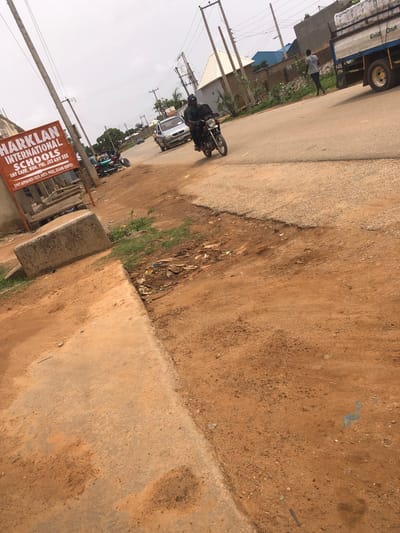 The image shows a street scene in Hanwa, Nigeria. A large truck is parked on the left side of the road, next to a man walking along the roadside. A motorcycle is also on the street. A sign for Harklan International Schools is visible near the bottom of the frame. The road is a mix of paved and unpaved surfaces, with dirt and debris along the edges. Overhead power lines are visible.
