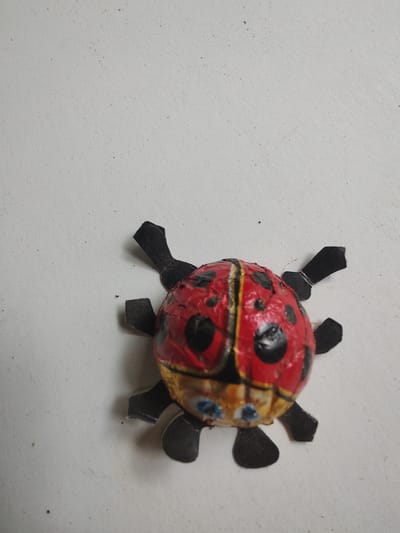 A whimsical, handmade ladybug figurine is the sole subject of this close-up, overhead shot. The ladybug is crafted from what appears to be crumpled and painted paper or foil, with a predominantly red body adorned with black spots. Yellow and black bands divide the body, and two small, blue circular "eyes" are visible on its head. Black paper strips form six legs, which are positioned as if the ladybug is crawling. The figurine is placed on a light-colored, textured surface, likely a wall or table, with small specks of dirt or debris scattered across it. The lighting is even, suggesting an indoor setting or an overcast day, with no discernible shadows. There is no visible text in the image. The overall impression is that of a charming, if slightly imperfect, craft project.