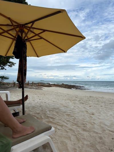 The image depicts a person's legs and feet resting on a lounge chair on a sandy beach, with a yellow umbrella providing shade. The scene looks out towards the ocean, with calm waves and a cloudy sky. The sand is rippled, suggesting it is soft and untouched by many footsteps. The atmosphere is relaxed and peaceful, as one would expect on a tropical beach. The time of day appears to be overcast, possibly late morning or afternoon, with diffused light. The presence of the lounge chair and umbrella indicates a resort or a designated relaxation area. The ocean appears to be a light blue-grey, and the horizon is clearly visible. Small rocks are scattered along the shoreline. In the foreground, the person's bare feet are prominent on the cushioned lounge chair. The overall impression is one of a tranquil beach experience.